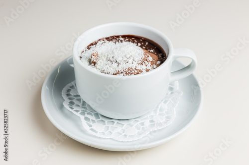 A small white cup of dark hot chocolate with chocolate ice-cream and coconut flakes on a white saucer and a napkin