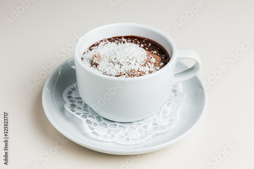 A small white cup of dark hot chocolate with chocolate ice-cream and coconut flakes on a white saucer and a napkin © jamurka