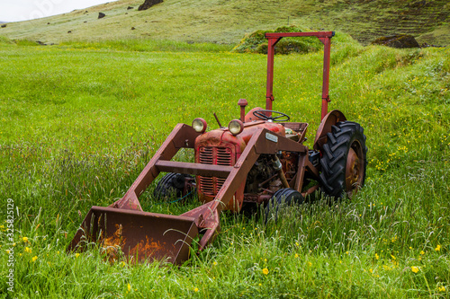 Old rusty red tractor in the middle of grassy meadow. Alone in the middle of nowhere. Agricultural machine with front facing plough. 