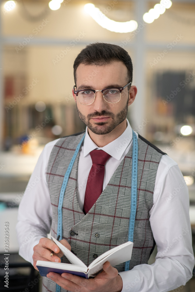 Male tailor standing, wearing glasses, holding notebook Male tailor standing, wearing glasses, holding notebook