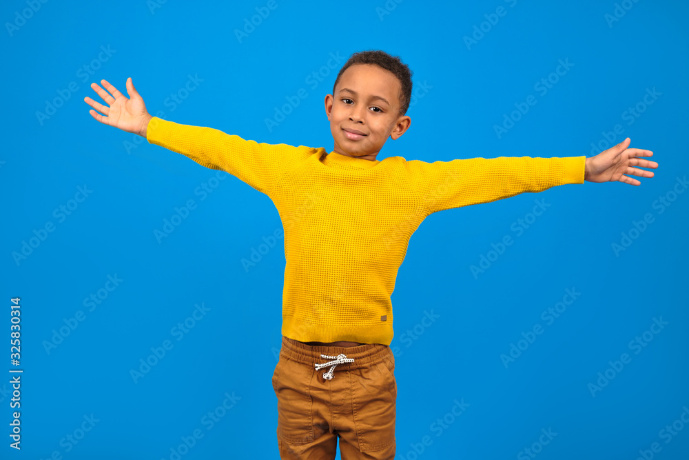 Happy African-American boy rejoices and smiles at victory or achievement, raises his hands up, on a blue studio homogeneous background. concept of winning, achieving the goal of striving to win.