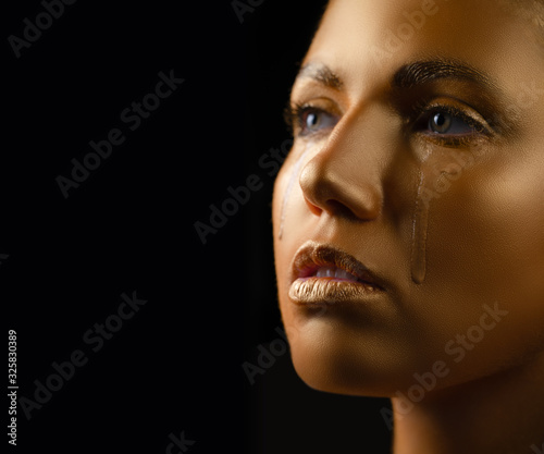 Beautiful adult girl in gold paint.Golden tears flowing from the eyes. Close-up portrait on a black background. Body art gold shiny paint. face shoulders and hair in makeup paint shine metallic