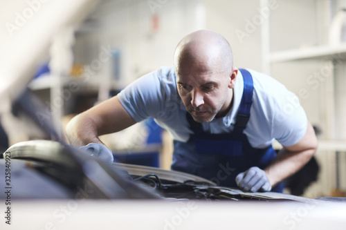 Front view portrait of muscular car mechanic looking into open hood of vehicle during inspection in garage shop, copy space