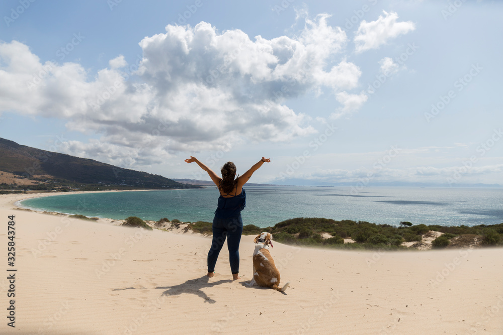 woman raising her arms happy with a large tan and white dog on top of a dune looking at the blue sea, green vegetation, windy and sunny day, amazing landscape