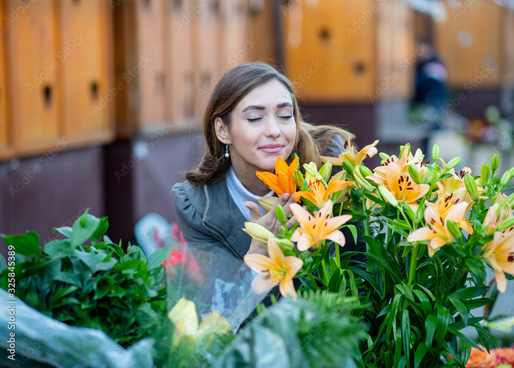 A woman smelling lilies in front of a flower shop on the street. She is adored by them.