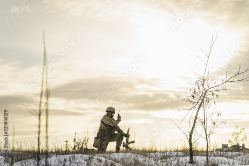 Modern war soldier army Man in camouflage speaks on the walkie-talkie radio crouching on one leg in the winter field. commandos with gun. soldier, sun backlight. modern warfare. atmosphere of battle