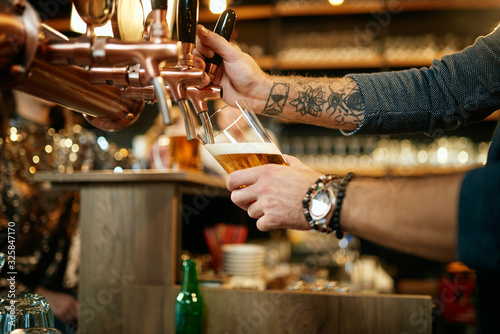 Tattooed caucasian barman pouring beer while standing in pub. Selective focus on hand.