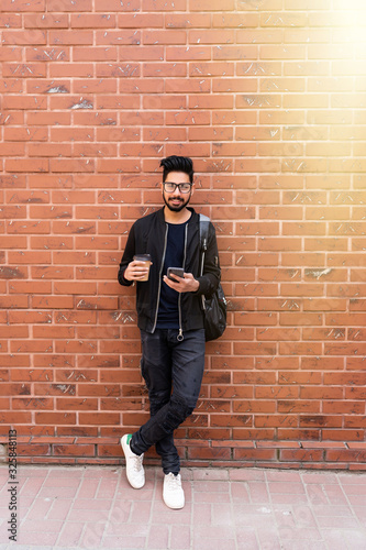 Young handsome indian man with phone and coffee cup standing against brick wall
