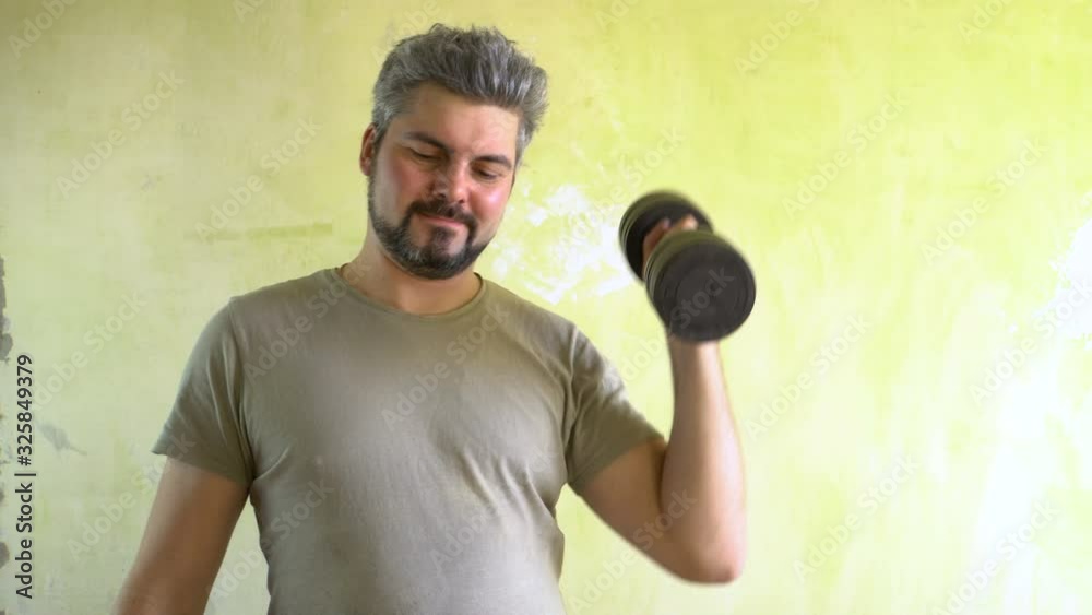A man with a beard is standing against the wall. Holds a dumbbell in his hand. He is engaged in fitness. A positive look. Joyful face. Military T-shirt. A man with a beard is standing against the wall. Holds a dumbbell in his hand. He is engaged in fitness. A positive look. Joyful face. Military T-shirt.