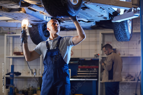 Portrait of muscular mechanic standing under car on lift while inspecting vehicle in auto repair workshop, copy space