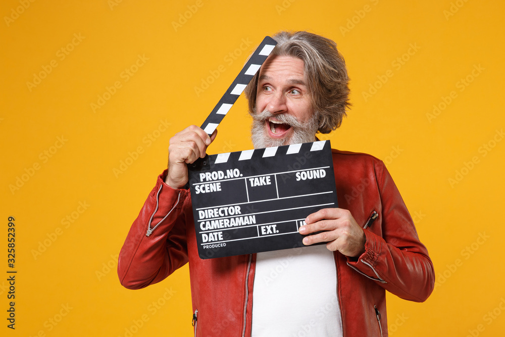 Amazed elderly gray-haired mustache bearded man in red leather jacket isolated on yellow orange background. People lifestyle concept. Mock up copy space. Hold classic black film making clapperboard.