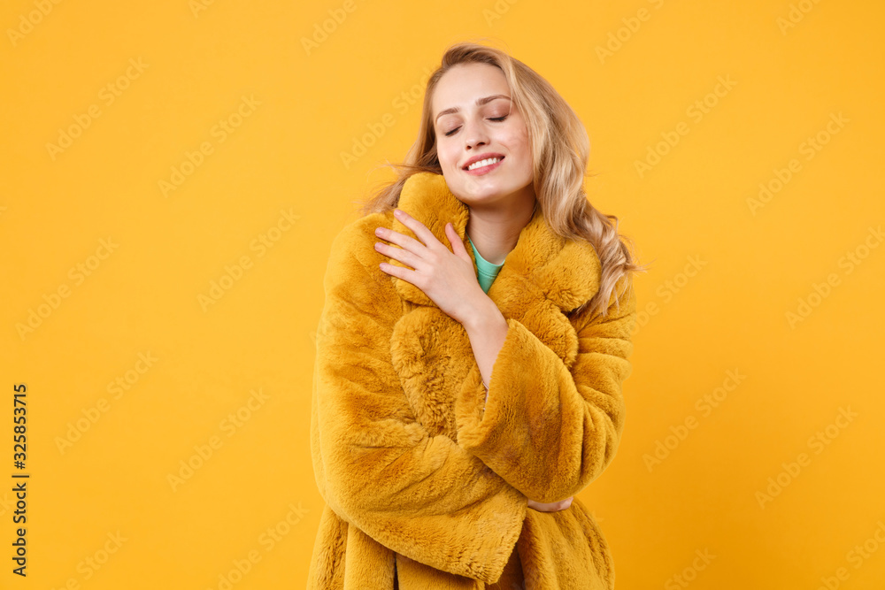 Smiling tender young blonde woman girl in yellow fur coat posing isolated on orange background, studio portrait. People lifestyle concept. Mock up copy space. Keeping eyes closed, hugging herself. Smiling tender young blonde woman girl in yellow fur coat posing isolated on orange background, studio portrait. People lifestyle concept. Mock up copy space. Keeping eyes closed, hugging herself.
