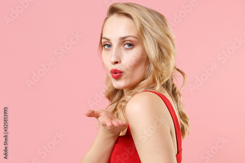 Side view of young blonde woman girl in red sexy clothes posing isolated on pastel pink background in studio. People lifestyle concept. Mock up copy space. Looking camera, blowing sending air kiss.