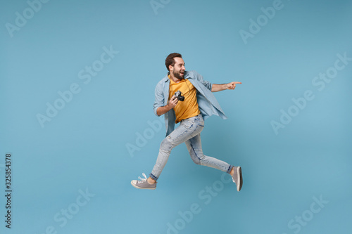 Excited traveler tourist man in yellow clothes with photo camera isolated on blue background. Male passenger traveling abroad on weekend. Air flight journey concept. Jumping, point index finger aside.