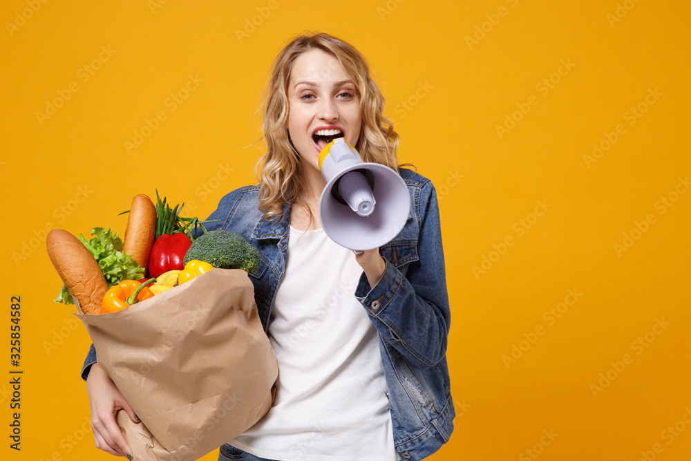Funny woman in denim clothes isolated on orange background. Delivery service from shop or restaurant concept. Hold brown craft paper bag for takeaway mock up with food products scream in megaphone. Funny woman in denim clothes isolated on orange background. Delivery service from shop or restaurant concept. Hold brown craft paper bag for takeaway mock up with food products scream in megaphone.