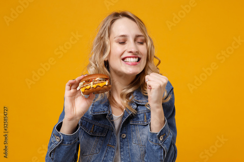 Happy young woman in denim clothes isolated on orange wall background. Proper nutrition or American classic fast food concept. Mock up copy space. Hold burger keeping eyes closed doing winner gesture.