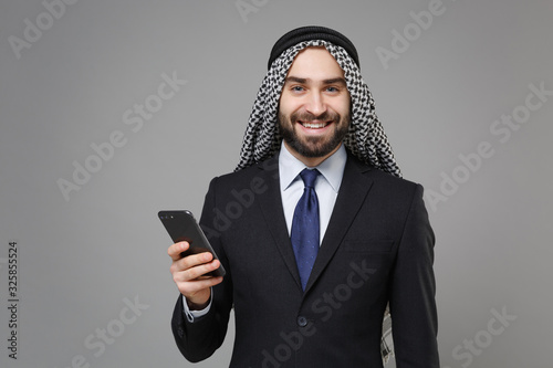 Smiling arabian muslim businessman in keffiyeh kafiya ring igal agal classic black suit isolated on gray background. Achievement career wealth business concept. Using mobile phone, typing sms message. photo