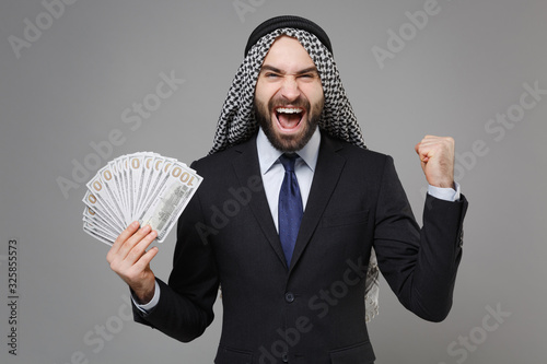Happy young arabian muslim businessman in keffiyeh kafiya ring igal agal black suit isolated on gray background. Achievement career wealth business concept Hold fan of cash money doing winner gesture. photo