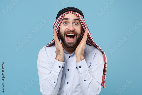 Surprised young bearded arabian muslim man in keffiyeh kafiya ring igal agal casual clothes isolated on pastel blue background. People religious lifestyle concept. Keeping mouth open, spreading hands. photo