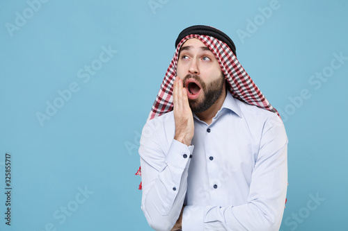 Tired young bearded boring arabian muslim man in keffiyeh kafiya ring igal agal casual clothes isolated on pastel blue background. People religious lifestyle concept. Yawning covering mouth with hand.