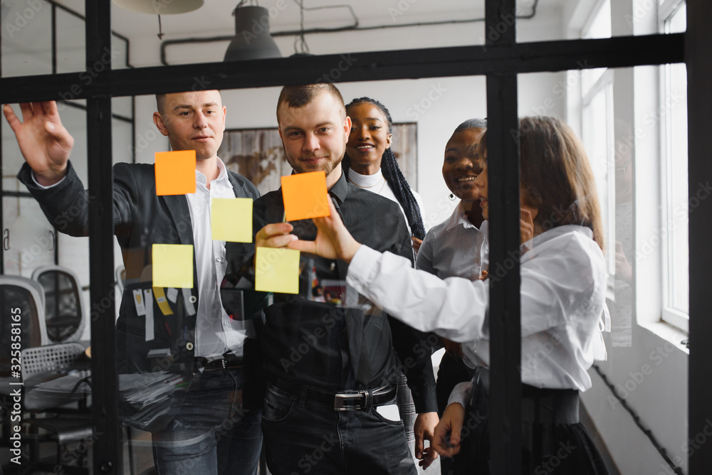 Happy mixed race colleagues watching smiling team leader marking task as done on colorful sticky notes of kanban agile glass window board at office. Diverse teammates satisfied with working process. Happy mixed race colleagues watching smiling team leader marking task as done on colorful sticky notes of kanban agile glass window board at office. Diverse teammates satisfied with working process.
