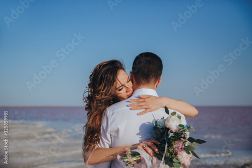 beautiful bride in a wedding dress on the seashore with a bouquet hugging her groom. Back view of the groom photo