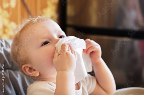 child wipes his nose with white handkerchief. sick child eats sitting on feeding chair. Kid with cold rhinitis. virus and infection. Coronavirus symptom