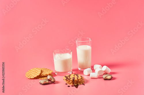 Two full glasses of fresh milk  yogurt or kefir near thin waffles  chocolate candies  marshmallow on pink background. Space for text