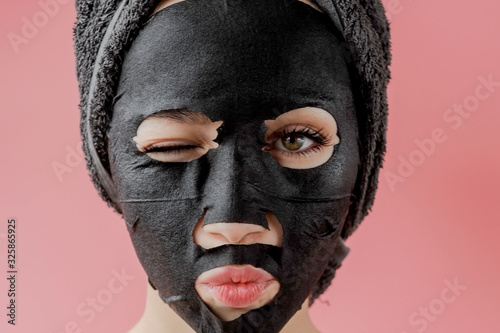 Young woman apply black cosmetic fabric facial mask on pink background. Face peeling mask with charcoal, spa beauty treatment, skincare, cosmetology. Close up