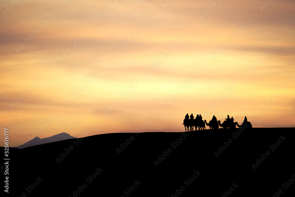 A group of camels and their riders arriving at the top of a sand dune are silhouetted against a dramatic cloudy desert sunset. A distant mountain peak rises above the horizon in the background.