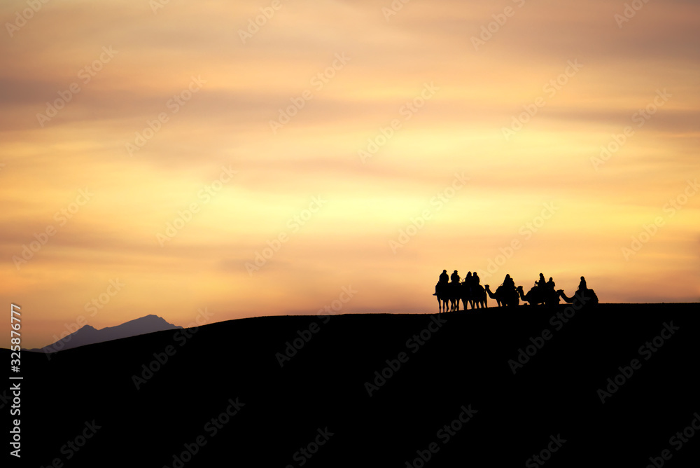 A group of camels and their riders arriving at the top of a sand dune are silhouetted against a dramatic cloudy desert sunset. A distant mountain peak rises above the horizon in the background. A group of camels and their riders arriving at the top of a sand dune are silhouetted against a dramatic cloudy desert sunset. A distant mountain peak rises above the horizon in the background.