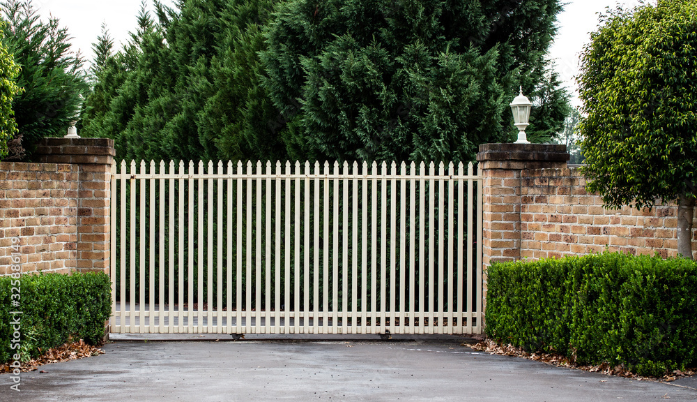Metal wrought iron driveway property entrance gates set in brick fence, garden trees in background Metal wrought iron driveway property entrance gates set in brick fence, garden trees in background
