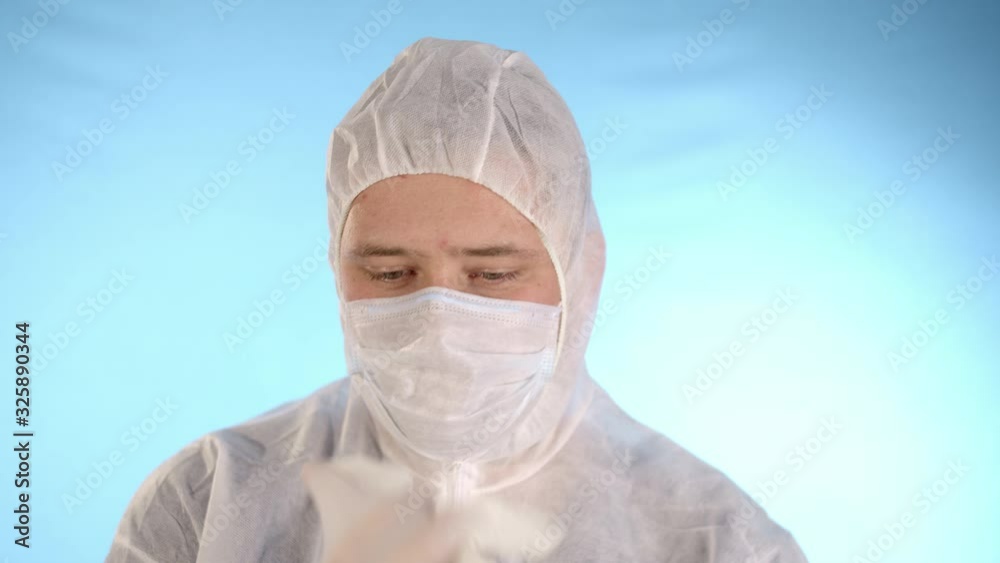 Caucasian man in a white protective suit and medical mask on a blue background puts rubber blue gloves on his hands Caucasian man in a white protective suit and medical mask on a blue background puts rubber blue gloves on his hands