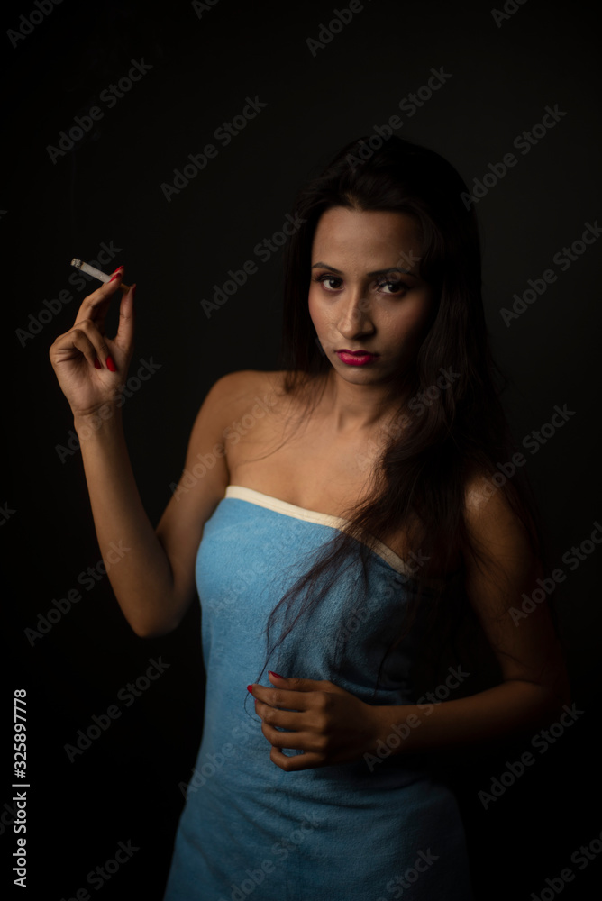 Fototapeta premium Portrait of Asian/Japanese/Korean/Indian brunette young girl in blue bath towel smoking cigarette in black studio copy space background. Indian lifestyle and fashion photography