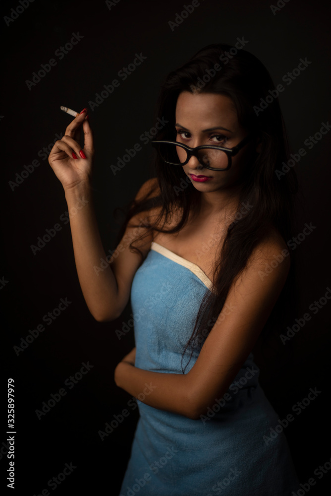 Fototapeta premium Portrait of Asian/Japanese/Korean/Indian brunette young girl in blue bath towel and spectacles smoking cigarette in black studio copy space background. Indian lifestyle and fashion photography