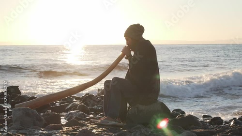 View of man sitting on seashore in sunlight and playing didjireedoo instrument photo