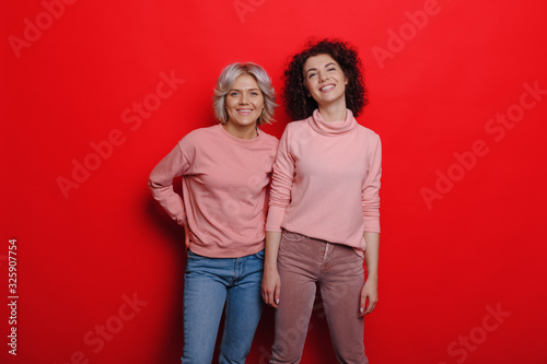 Two curly haired confident caucasian women posing in sweaters on red background © Strelciuc