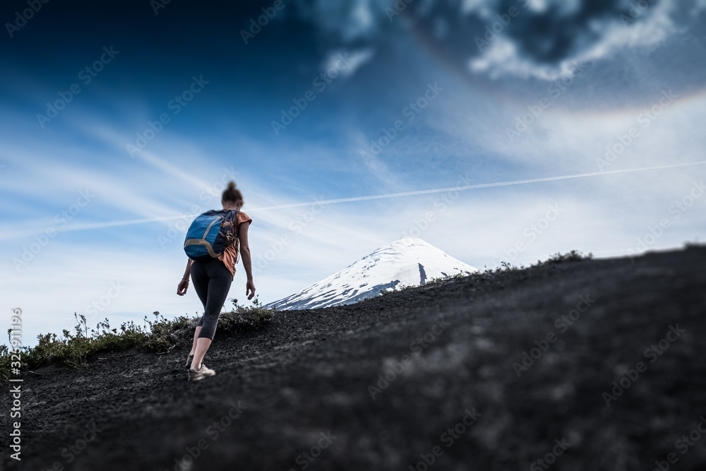 Young woman hiker walks on the deserted loose rocky trail on the mountain slope with snow capped volcano of Osorno on the background in Chile. Tilt shift effect applied
