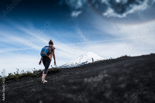 Young woman hiker walks on the deserted loose rocky trail on the mountain slope with snow capped volcano of Osorno on the background in Chile. Tilt shift effect applied