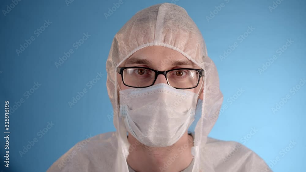 Long hair caucasian bearded man wearing glasses in a white protective jumpsuit on a blue background puts a medical mask on his face, zips his jacket and puts on a hood. Long hair caucasian bearded man wearing glasses in a white protective jumpsuit on a blue background puts a medical mask on his face, zips his jacket and puts on a hood.