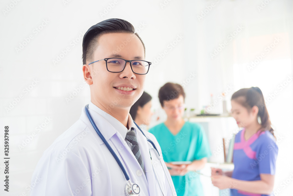 Young asian doctor in white coat smiling at camera with his team standing in background. Young asian doctor in white coat smiling at camera with his team standing in background.