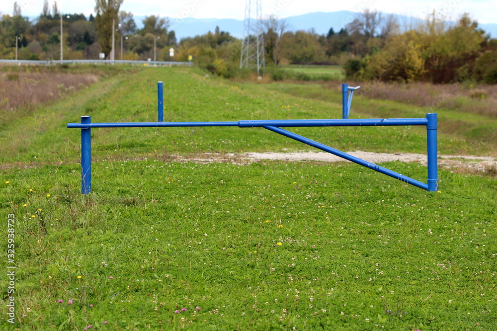 Closed and open strong blue metal ramps put around gravel path to prevent vehicle access to grass covered abandoned road surrounded with tall dry grass and trees Closed and open strong blue metal ramps put around gravel path to prevent vehicle access to grass covered abandoned road surrounded with tall dry grass and trees