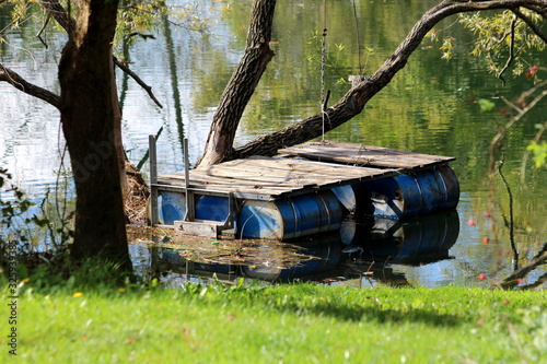Dilapidated homemade river platform made from blue metal barrels and wooden boards fixed to old large tree surrounded with calm river water and green grass covered river bank
