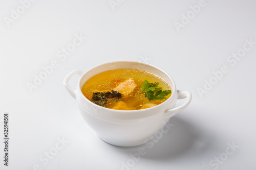 Fish soup with salmon. A white round plate is filled with a transparent broth, which shows pieces of salmon, potatoes and greens. Plate on a white background. Side view