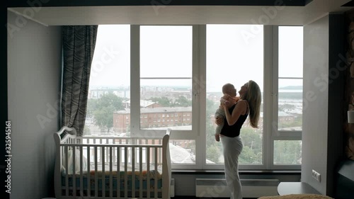 Young mother tossing her baby near the window. Happy blond woman holding her little son and playing with him near the beddy bye in the childroom with panoramic city view. photo