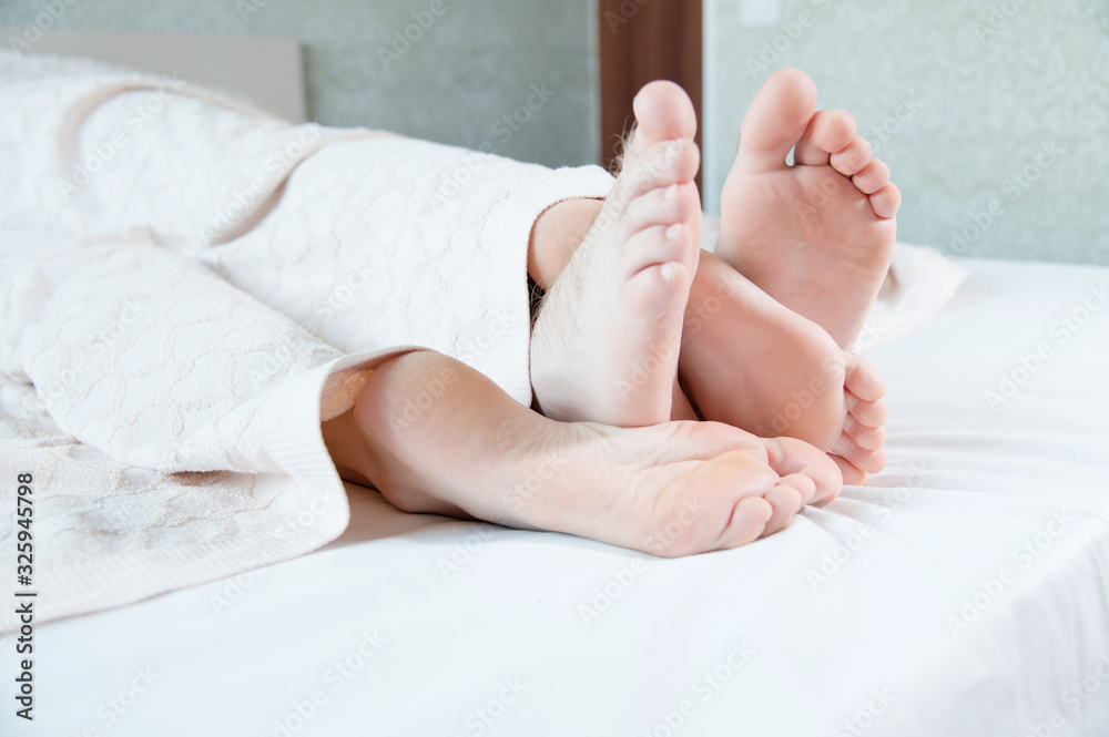 Close-up of the feet of a young couple sticking out from under the covers in the bedroom. Bare feet caress each other engaged in grooming. Close-up of the feet of a young couple sticking out from under the covers in the bedroom. Bare feet caress each other engaged in grooming.