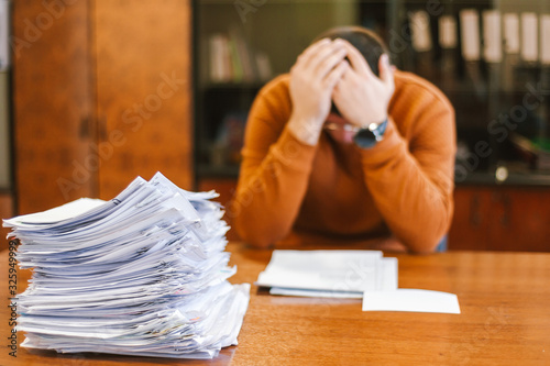 A businessman grabs his head from a pile of papers at work. The concept of depression from difficulties and possible bankruptcy of an enterprise.