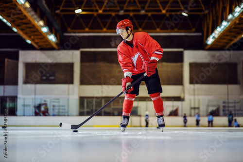 Full length of handsome hockey player standing on the ice in hall and preparing to make a score.