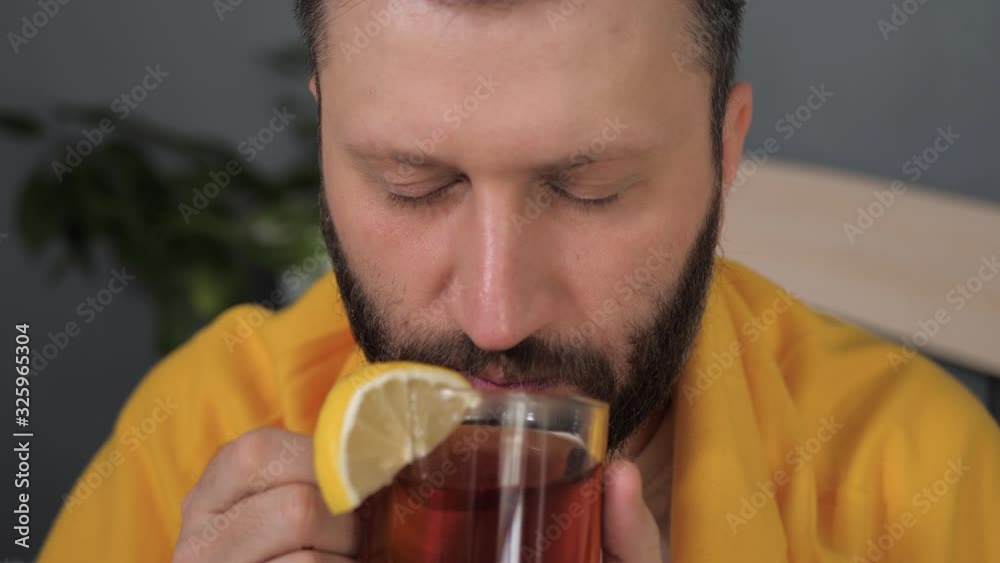 Front view guy is drinking black tea. Attractive young man is ill and drinks tea with lemon while sitting in bed. Cold, flu, sore throat, runny nose, acute respiratory disease concept. Slow motion Front view guy is drinking black tea. Attractive young man is ill and drinks tea with lemon while sitting in bed. Cold, flu, sore throat, runny nose, acute respiratory disease concept. Slow motion