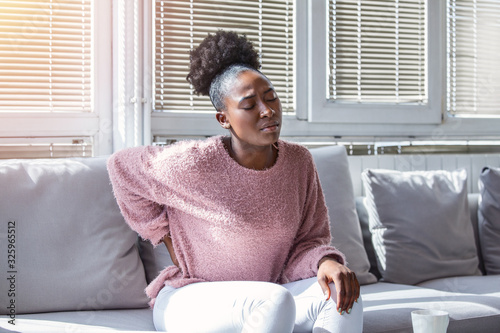 Young Black woman suffering from backache at home. Portrait of a young brunette girl sitting on the couch at home with a headache and back pain. Beautiful woman Having Spinal Or Kidney Pain photo